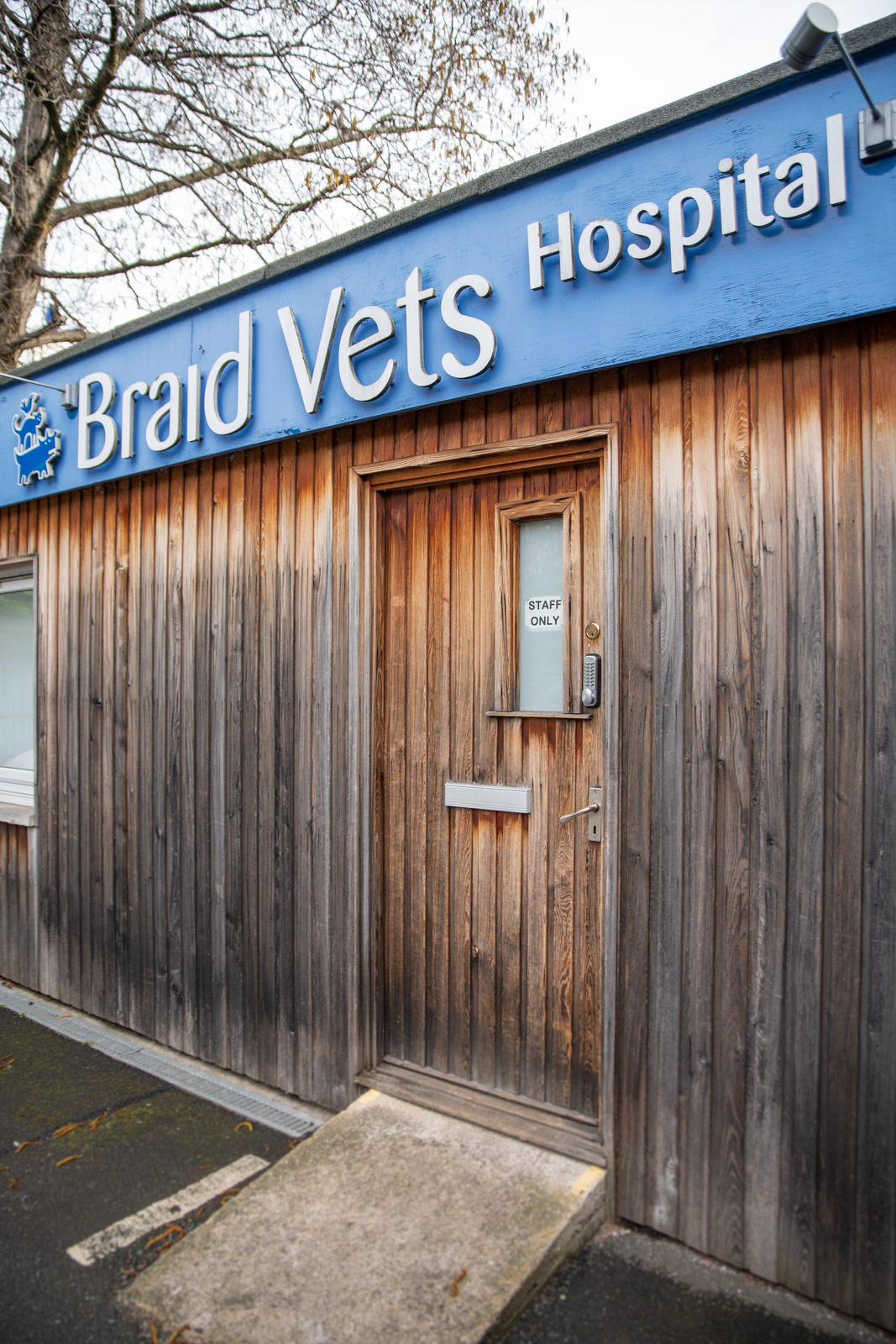 Wooden exterior of Braid Vets Hospital with a Staff Only door. Sign is blue with white lettering. Tree branches visible above the roof.