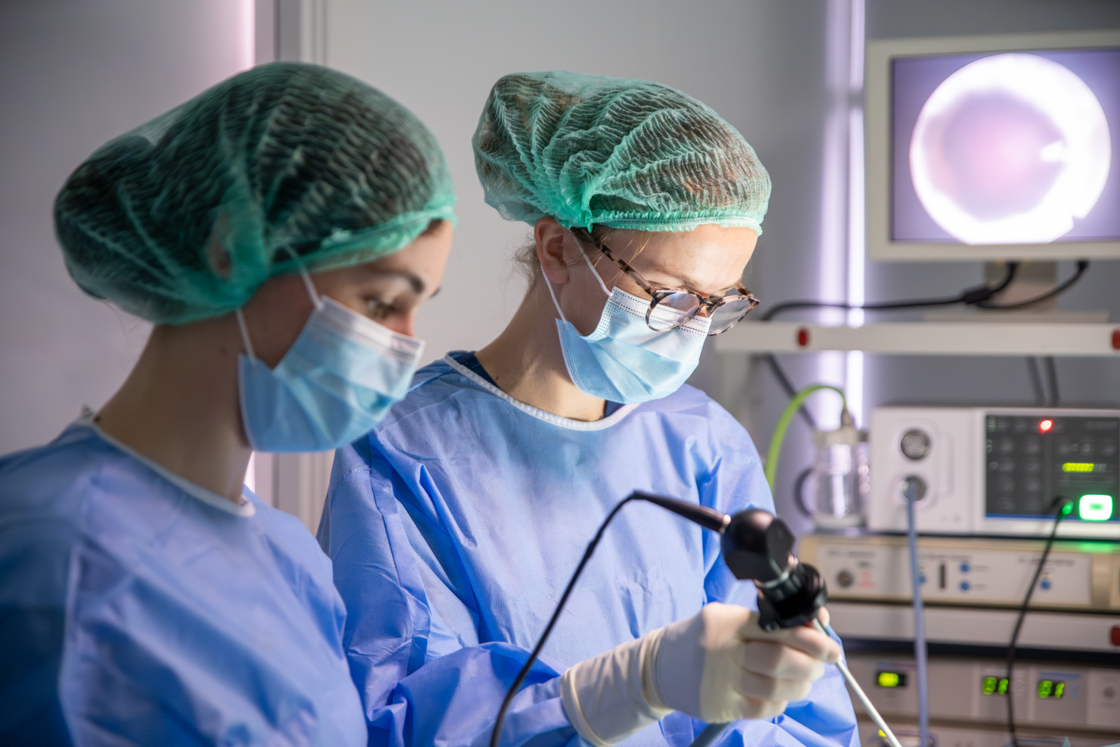 Two medical professionals wearing blue scrubs, masks, and hairnets perform a surgical procedure using endoscopic equipment. One is holding a camera, while the other observes. A monitor and medical devices are visible in the background.