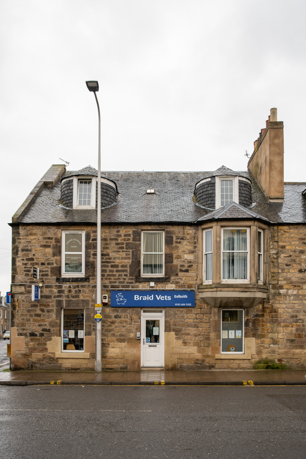A stone building with a sign that reads Braid Vets. It has two dormer windows on the roof and a shopfront with large windows. A streetlight stands on the sidewalk nearby under a cloudy sky.