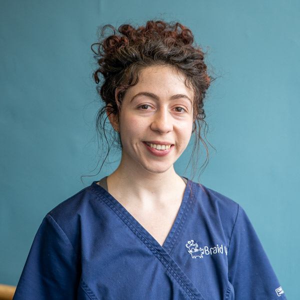 A smiling person with curly hair wearing a navy blue scrub top stands in front of a teal backdrop. Visible text on the scrub includes a logo and the word Braids.