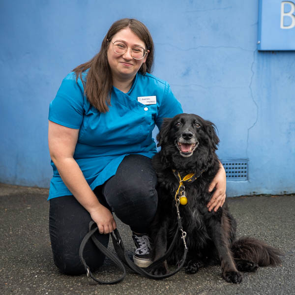 A woman in a blue uniform crouches beside a black dog with a yellow leash, both smiling. They are outdoors in front of a blue wall.