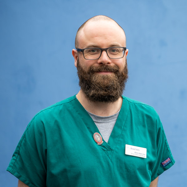 A person with a beard and glasses wearing green medical scrubs and a nametag stands against a blue background.