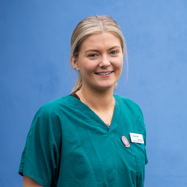 A person with blonde hair is smiling and wearing green medical scrubs with a name tag and a pin. They stand in front of a blue background.