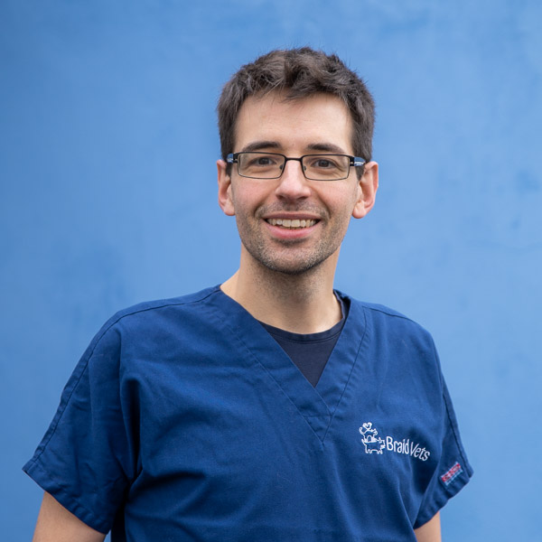 A person wearing blue scrubs with Braid Vets embroidered on the top stands against a blue background. They have glasses and short dark hair, smiling at the camera.