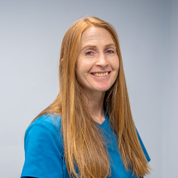 A person with long red hair smiles and wears a blue shirt. The background is a plain off-white color.