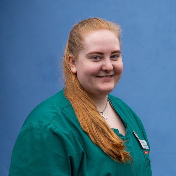 A person with long red hair wearing green medical scrubs smiles at the camera against a blue background. They have a name badge and a necklace.