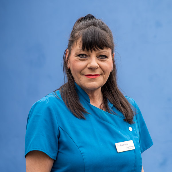 A woman with dark hair in a ponytail stands in front of a blue background, wearing a blue uniform and a name tag. She has a neutral expression and red lipstick.