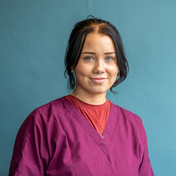 A person with dark hair is smiling, wearing a purple scrub top over a red shirt against a light blue background.