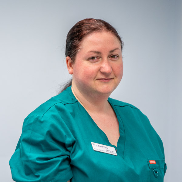 Person with dark hair tied back, wearing teal medical scrubs and a name tag, stands against a light gray background. They have a neutral expression.
